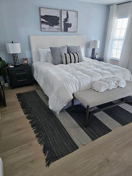 Bedroom with a white bed, gray bench, black and gray rug, two nightstands, and light blue walls.