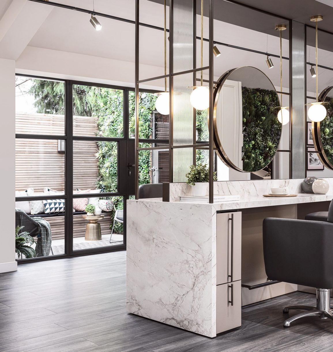 Modern salon interior with marble styling stations, round mirrors, pendant lights, and large windows.