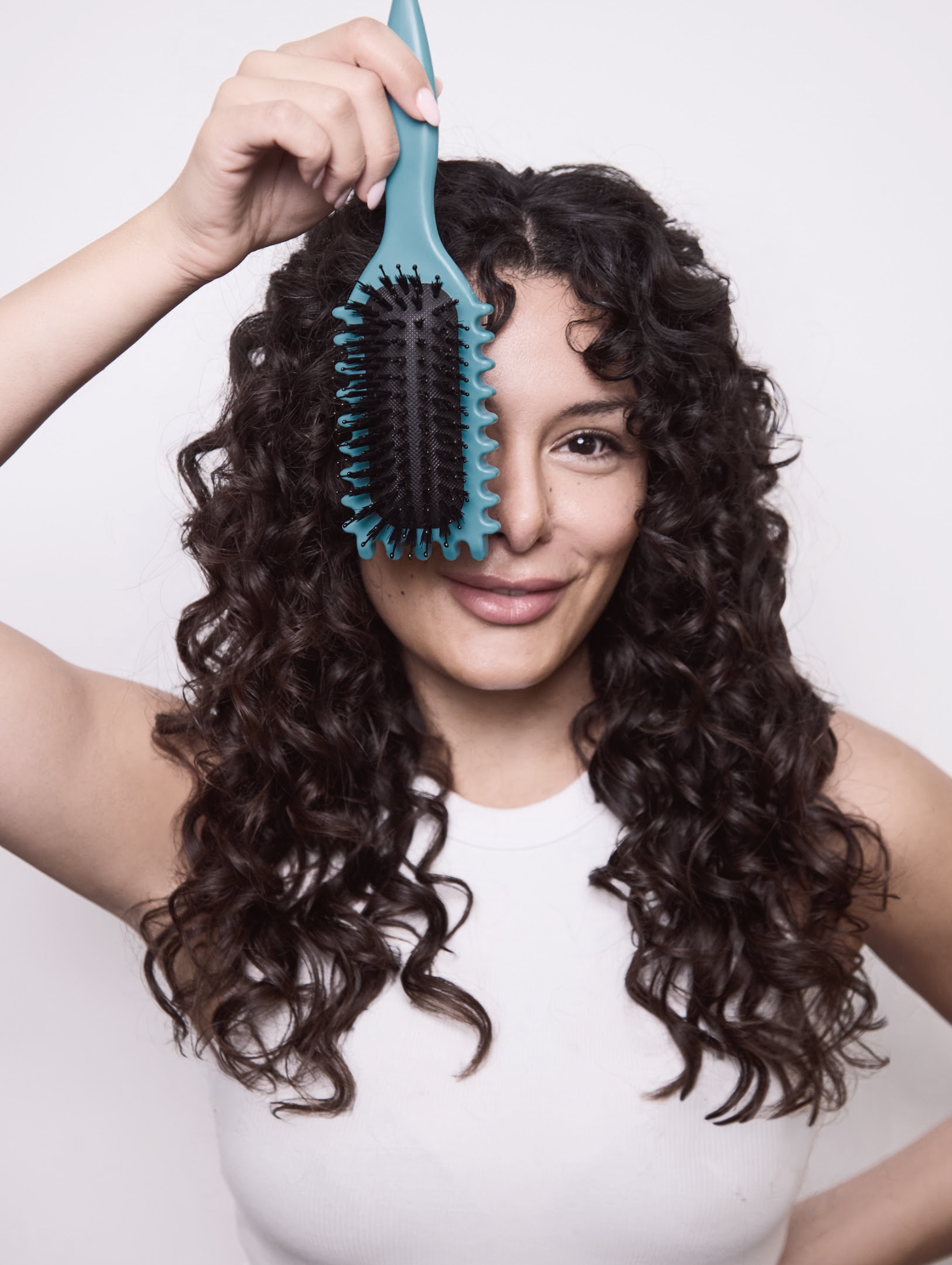 Leo Bancroft model with curly hair holding a turquoise hairbrush over her face, smiling against a light background