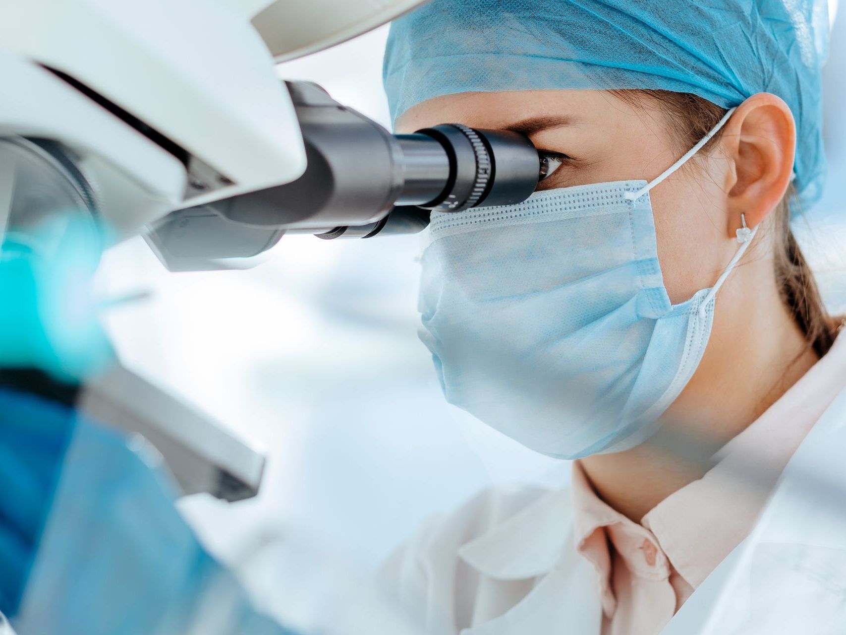 A woman wearing a mask is looking through a microscope.