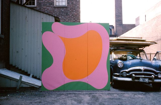 A blue car is parked in front of a pink and orange painting