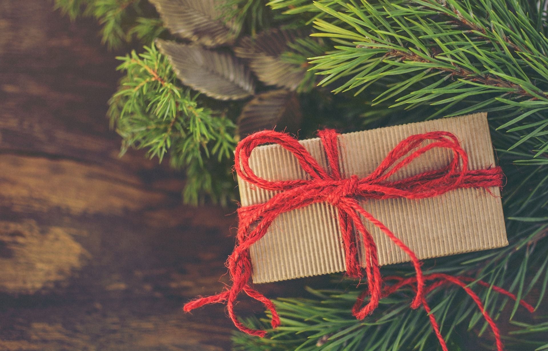 Gift box wrapped in brown paper with a red twine bow, nestled with evergreen branches on a wood surface.