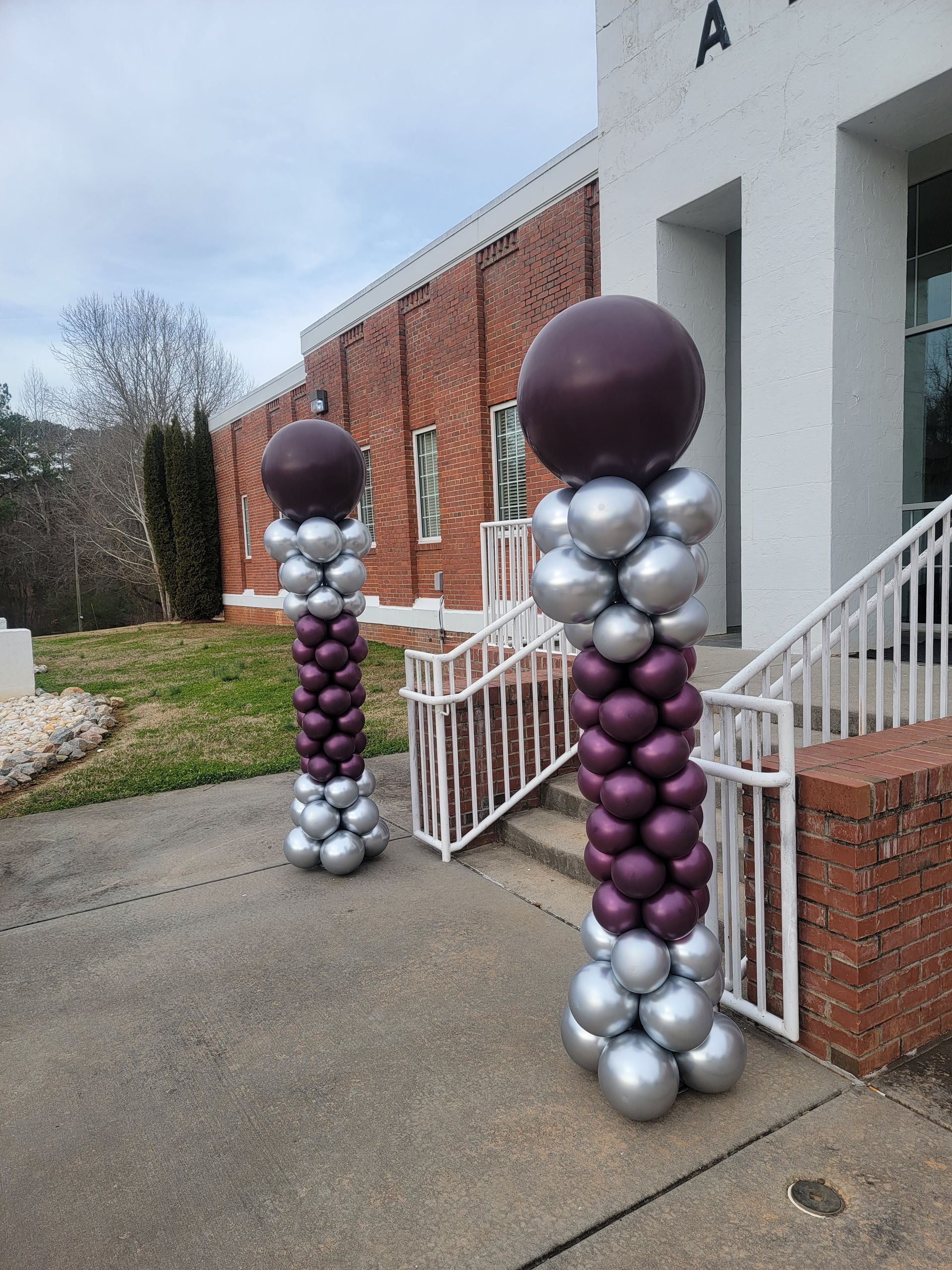 Two balloon columns, silver and purple, outside a brick building with white stairs.