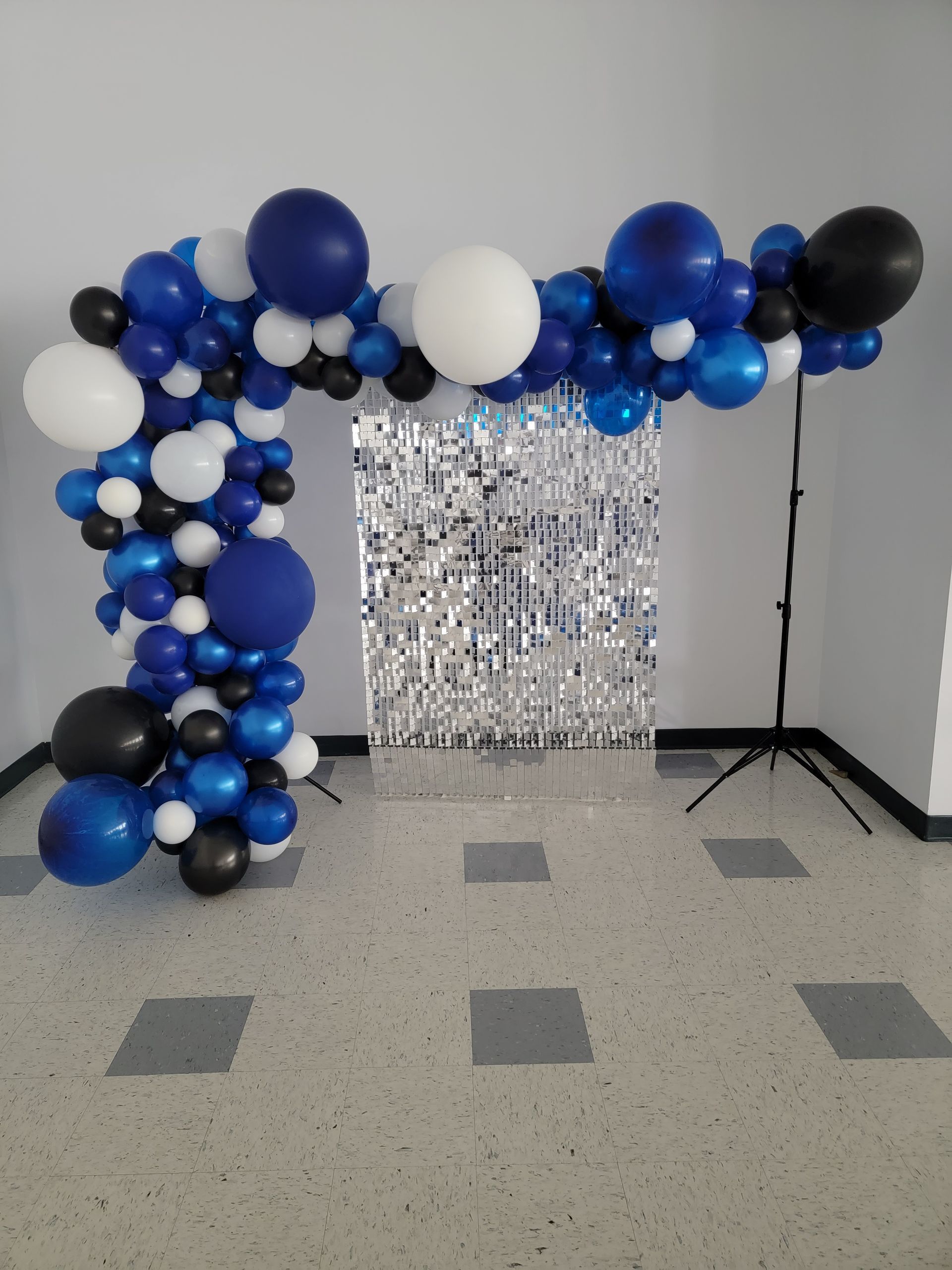Blue, white, and black balloon arch over a silver fringed backdrop. Setting: indoors, floor with square tiles.