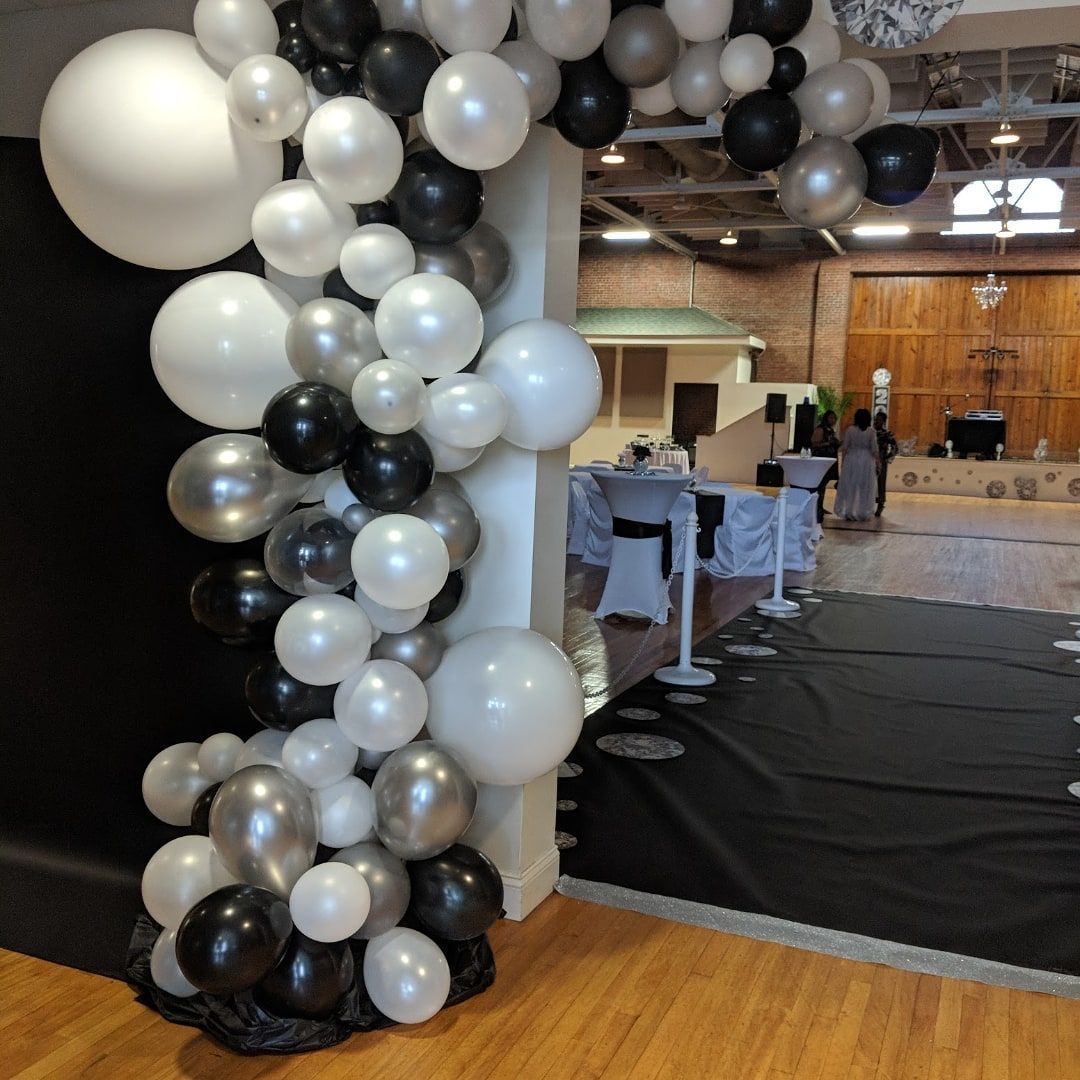 Black, white, and silver balloon arch at a formal event entrance; black carpet and white table linens.
