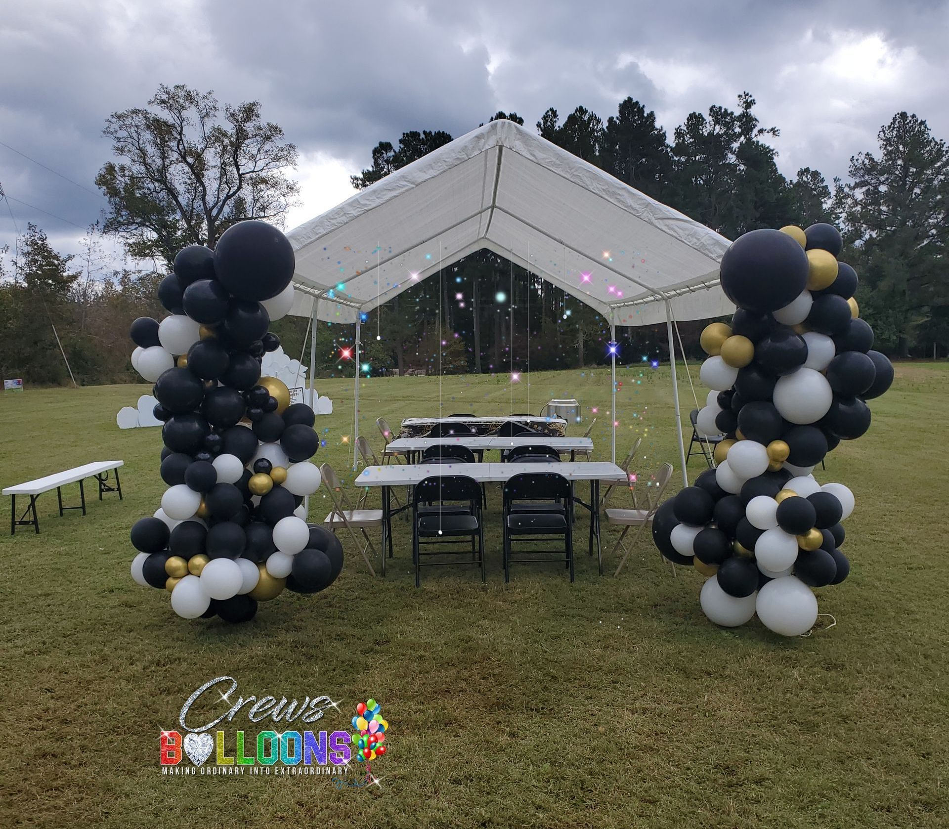 Party setup with a tent, tables, chairs, and balloon decorations in black, white, and gold, outdoors.