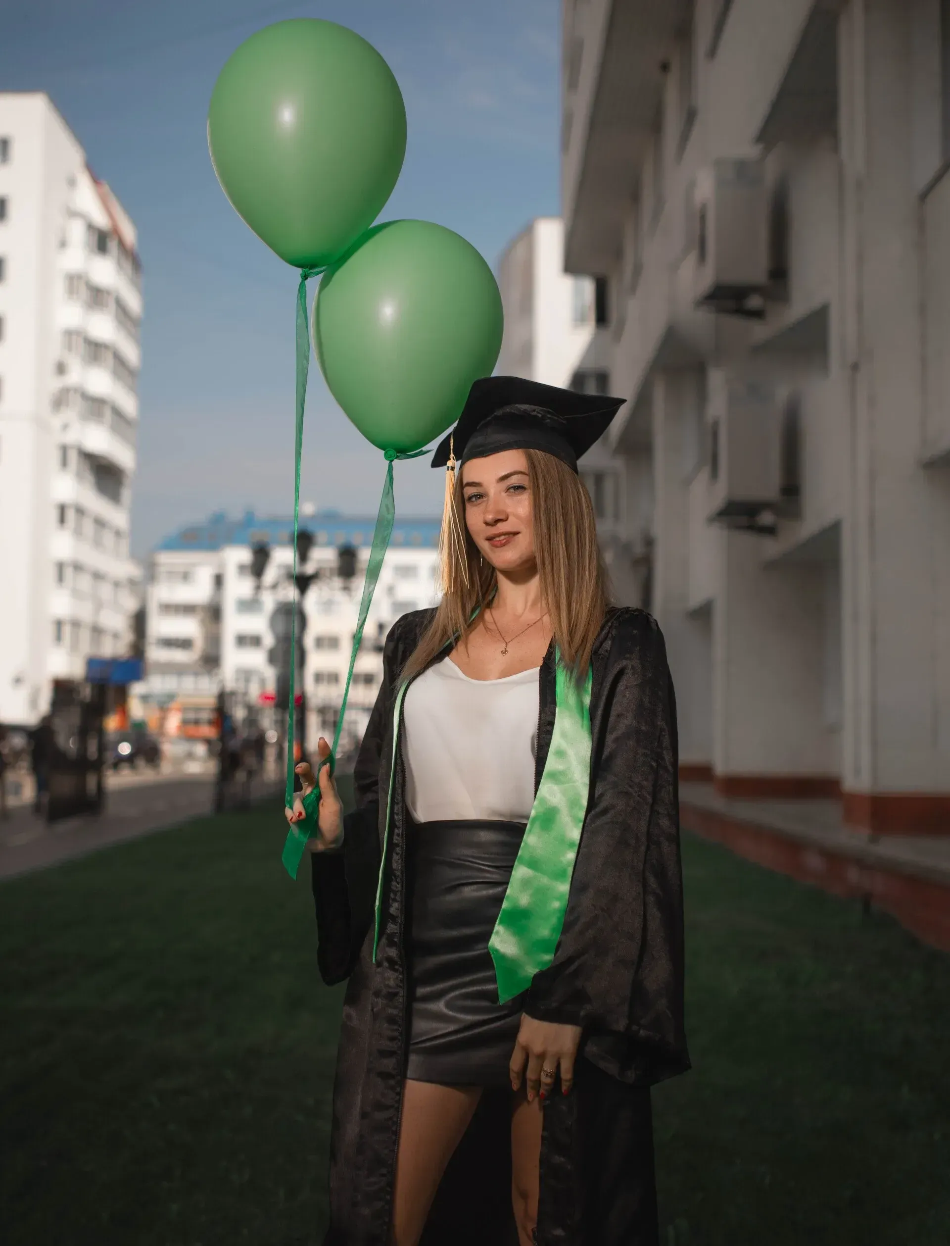Woman in graduation cap and gown holding green balloons outdoors.