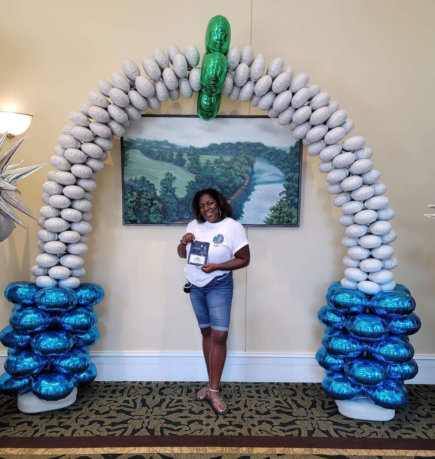 Woman holding a book stands under a blue and silver balloon arch in a decorated room.