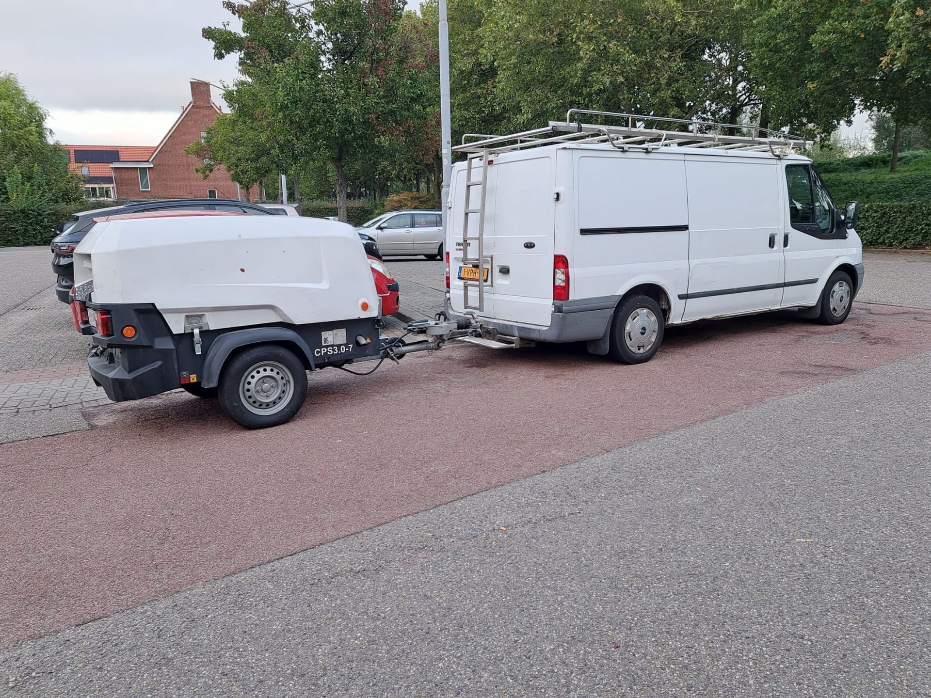 Op een parkeerplaats staat een wit busje met een aanhangwagen eraan vast.