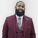 A man with a beard is wearing a maroon suit and tie.