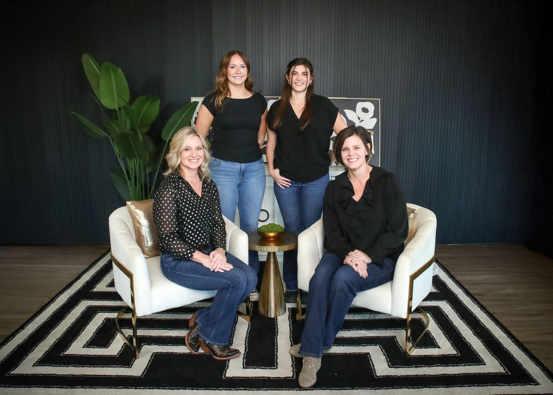 Four women posing in a modern room, two seated in white chairs, black and white rug, and large plant.