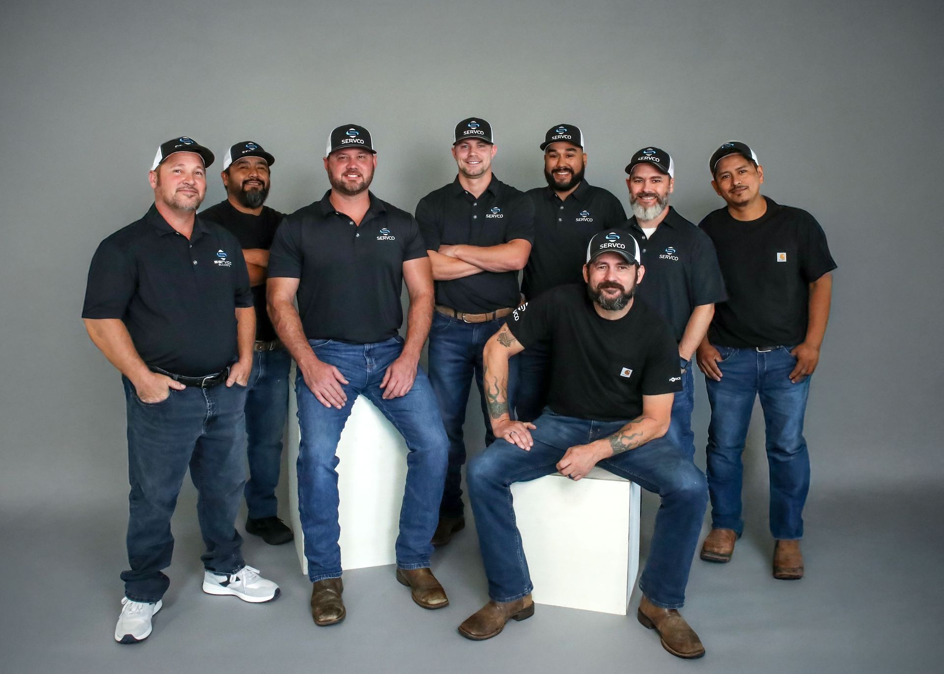 Group of men in black shirts and jeans, smiling, posing in front of a gray backdrop.