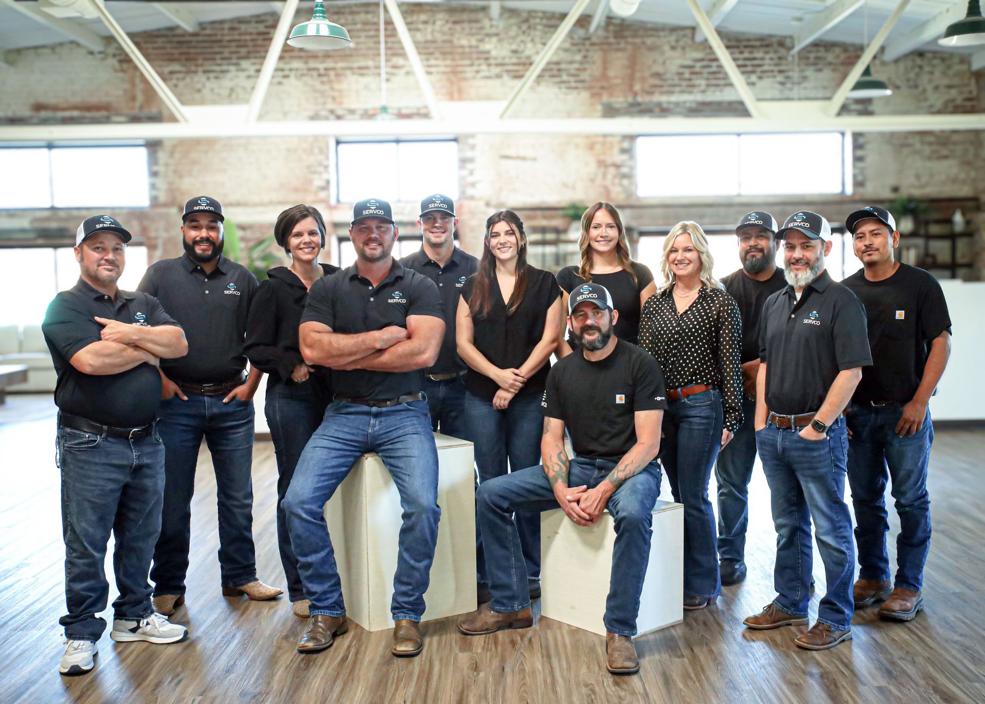 Group of people in black shirts and jeans pose in an office. Some have arms crossed, while others are smiling.