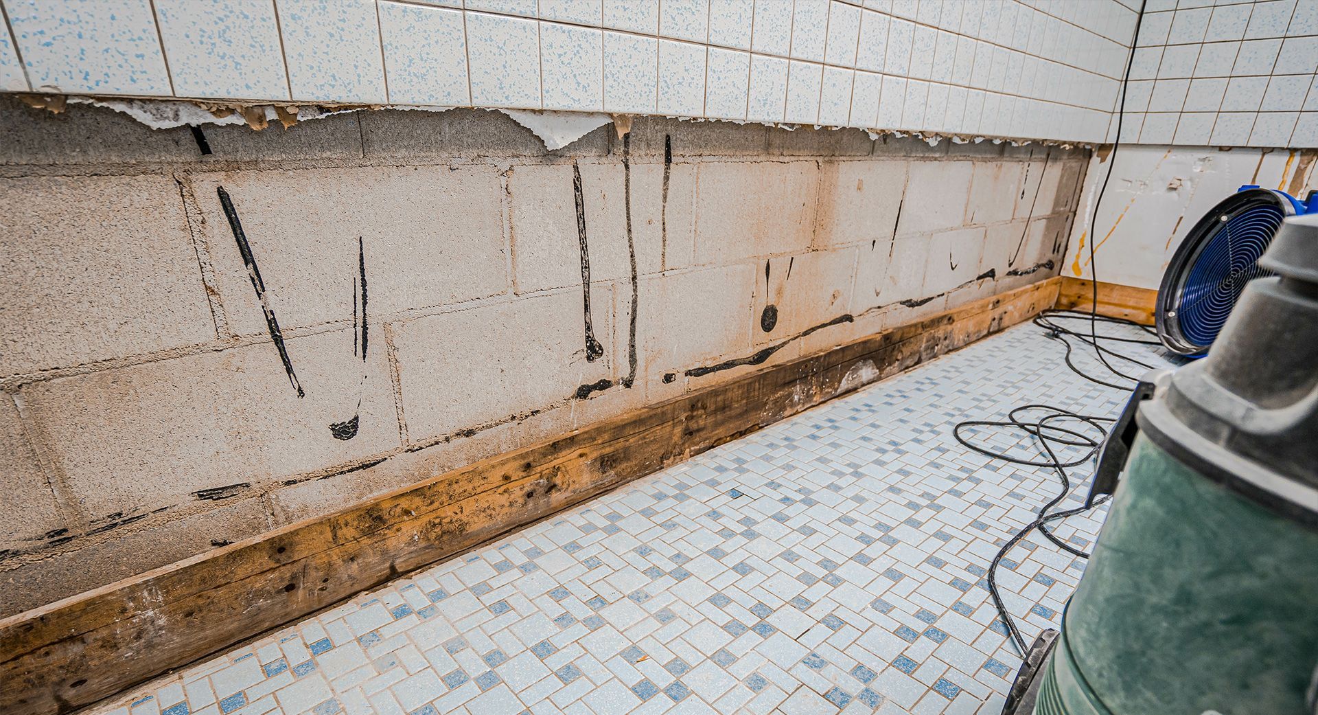 Damaged wall in bathroom, tiles partially removed, showing concrete blocks and water damage.