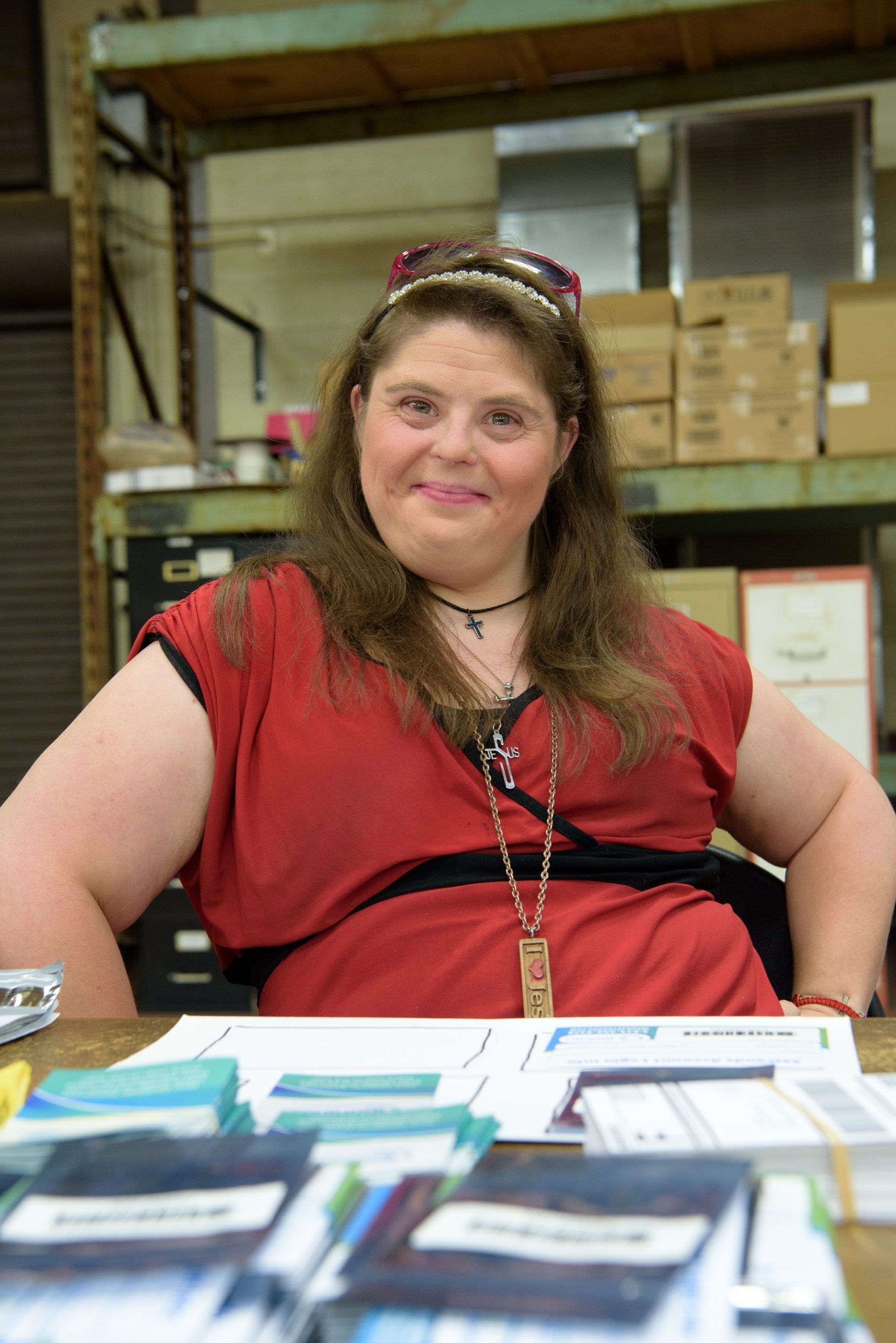 A woman in a red shirt is sitting at a desk in a warehouse.