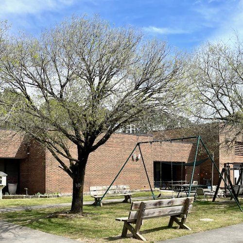 A park with a bench , swings , and trees in front of a brick building.