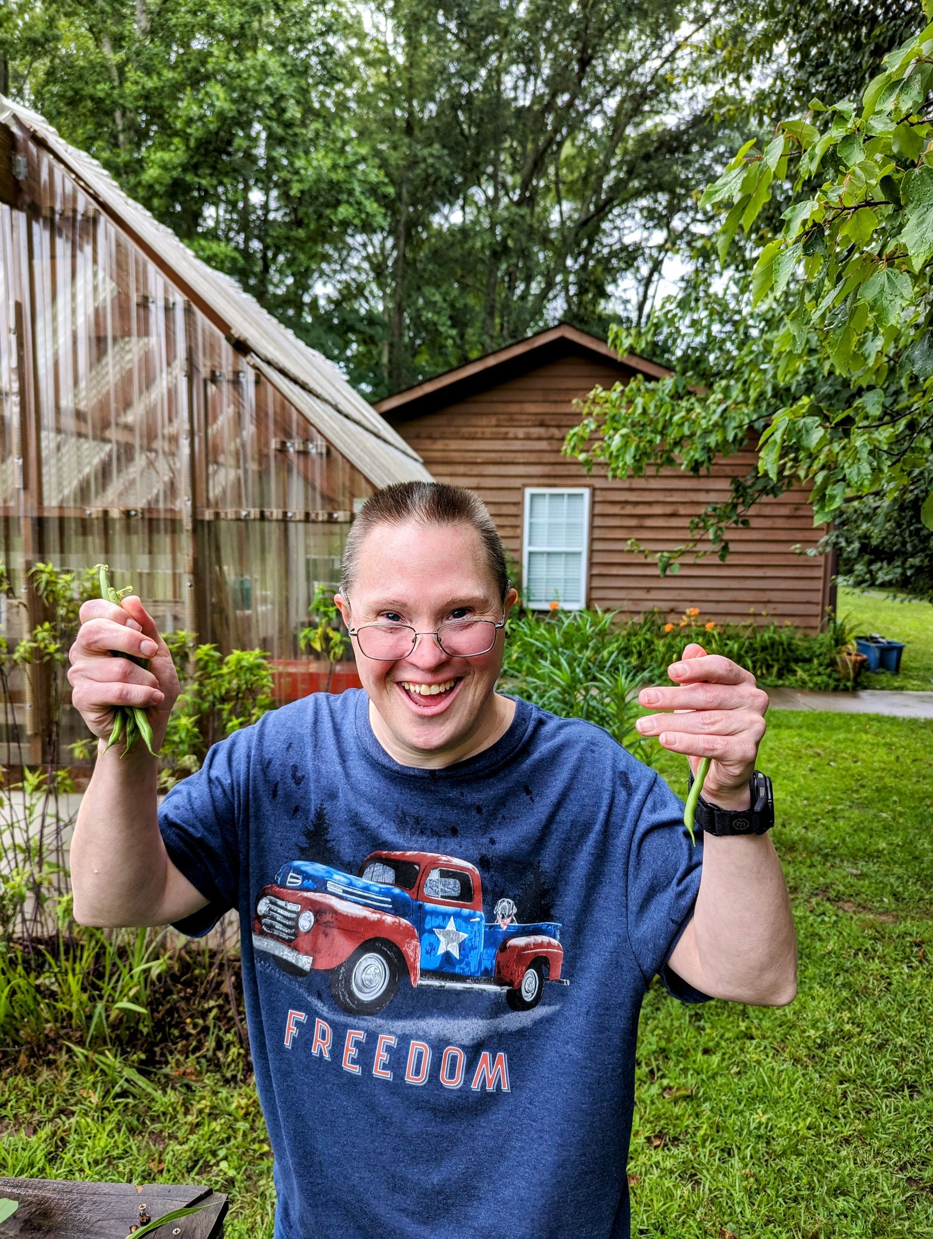 A man wearing a blue shirt with a truck on it is holding a plant in front of a house.