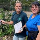 Two women are standing next to each other in a garden and one is holding a clipboard.