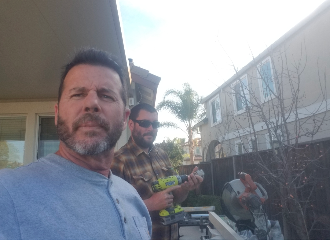 Two men work on an outdoor project, one holding a power drill, in a residential backyard with a house in the background.