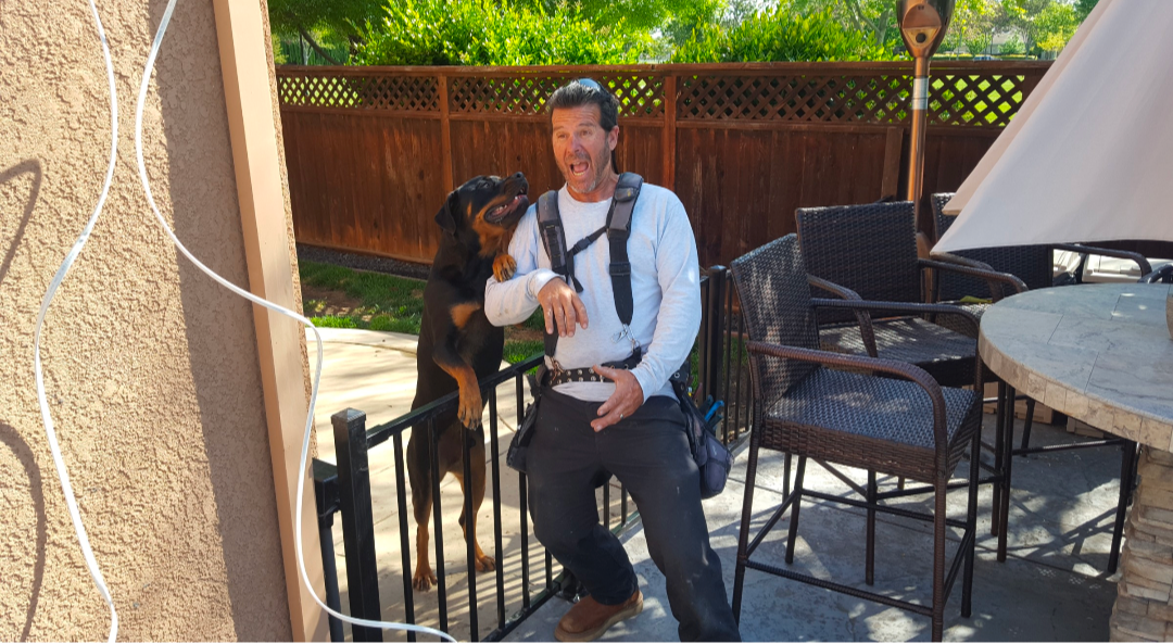 A person standing on a patio wearing a backpack harness as a dog stands on its hind legs to greet them.