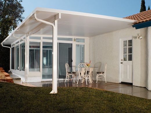 A covered white patio featuring a dining set, glass walls, and a white door attached to a house with a textured wall.
