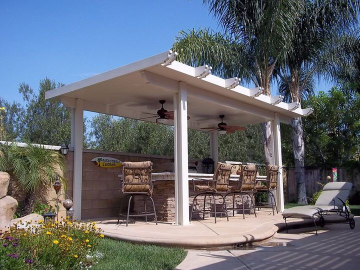 A covered outdoor patio with a stone bar, bar stools, and two ceiling fans, set in a sunny backyard with foliage.