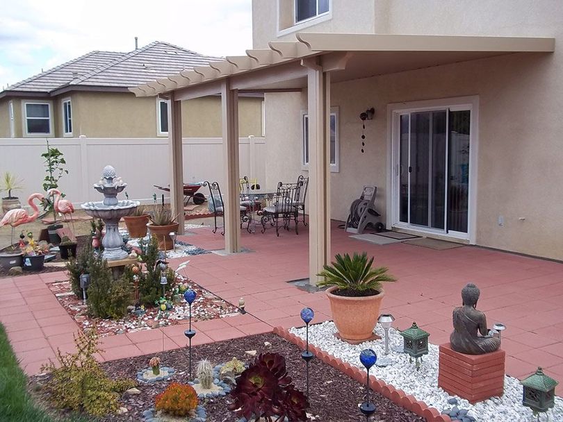 A residential patio with a shaded pergola, red pavers, a decorative fountain, potted plants, and garden ornaments.