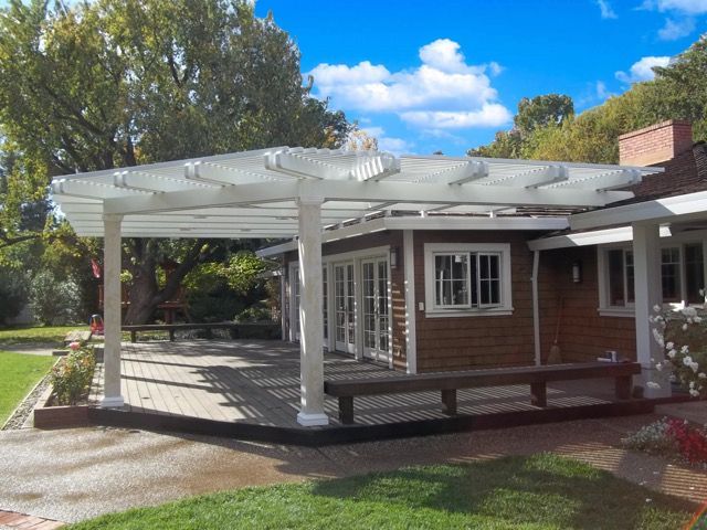 A white wooden pergola extends from a brown house over a paved patio, featuring a built-in wooden bench and garden area.