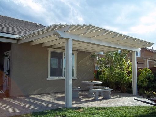 A white pergola shades a stone patio with a matching concrete table and bench set next to a beige house exterior.