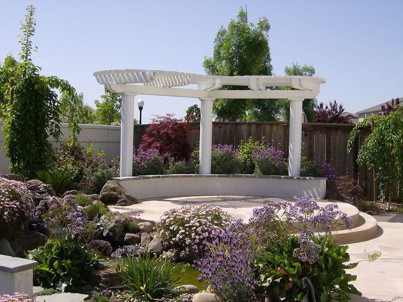 A backyard garden with a white pergola over a stone bench, lush green plants, purple flowers, and a small rock pond.