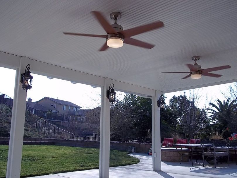 A covered outdoor patio featuring two ceiling fans with lights, white pillars, and wall-mounted lantern-style fixtures.
