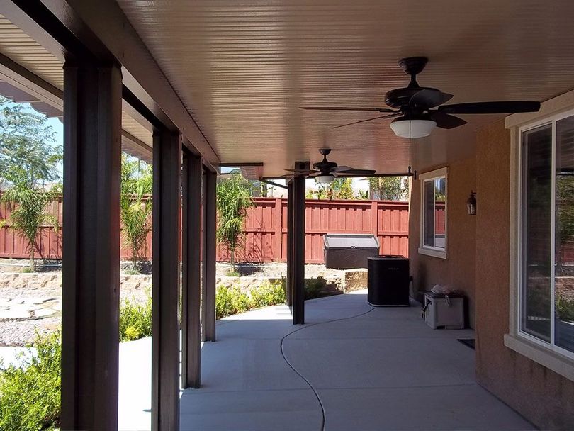 A covered patio with a concrete floor, wooden posts, two ceiling fans, and a view of a backyard with a red fence.