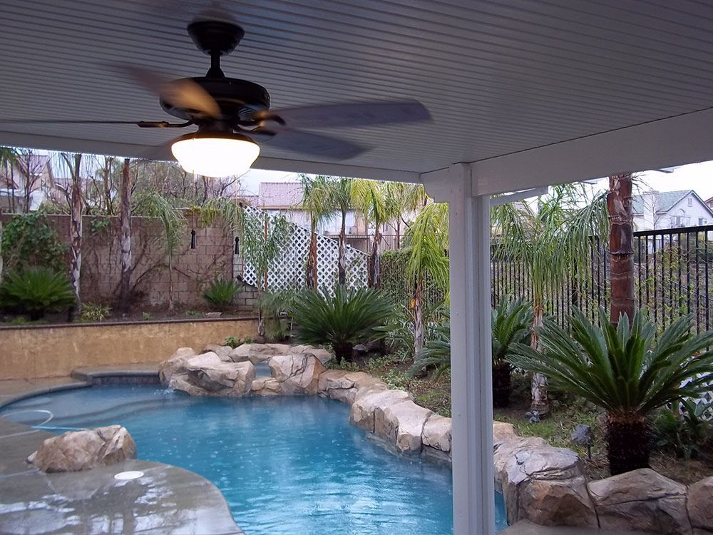 A swimming pool with rock features and lush landscaping, viewed from a covered patio with a ceiling fan.
