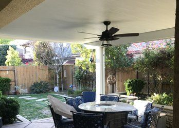 A patio dining set with a round table and blue chairs sits under a covered roof with a ceiling fan in a backyard.