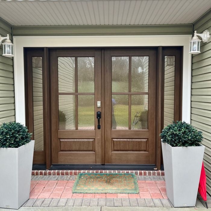 Brown double doors with glass panes, flanked by sidelights, surrounded by green siding and white trim. Two gray planters with green shrubs sit on brick.