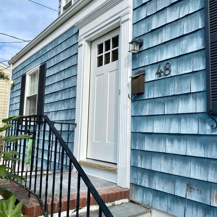 Blue-shingled house with white door, 48 house number, black shutters, and a wrought-iron railing.