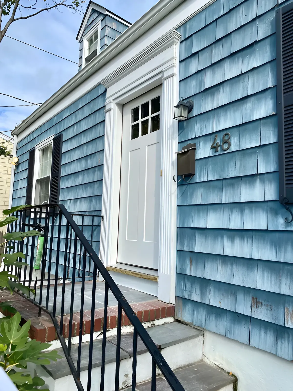 Blue-shingled house with white door, black shutters, and black iron railing. Number 49 on the wall.