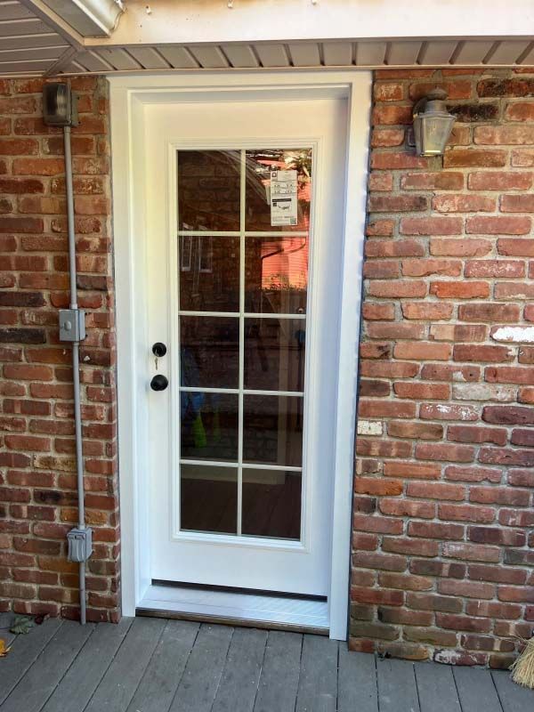 White-framed door with glass panes, set in a brick wall, next to a gray conduit, on a wooden deck.