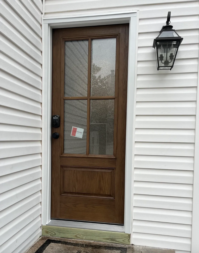 Brown wooden door with glass panels, mounted on white siding; black lock, and outdoor light fixture.