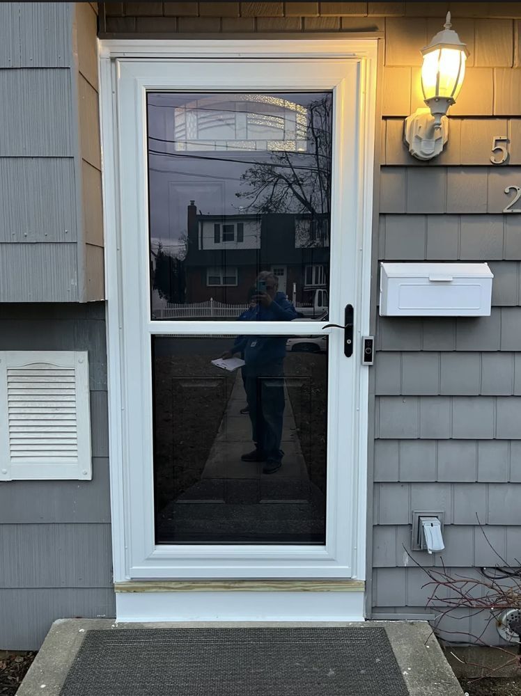 White front door with screen, mailbox, and light on gray siding; a person is visible through the door.
