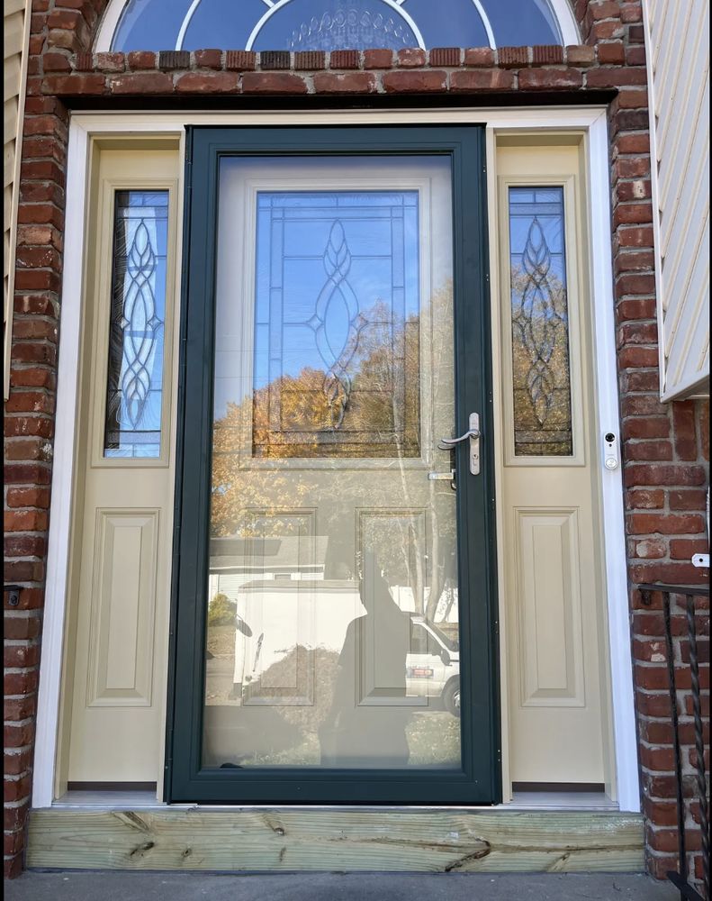 Front door with storm door, sidelights, and a brick facade. The door frame is painted green, the door and sidelights are beige.