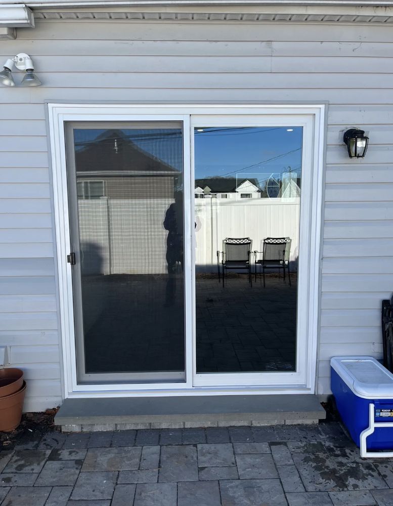 Sliding glass door in a grey-sided building with a stone patio, reflected sunlight, and two chairs visible outside.