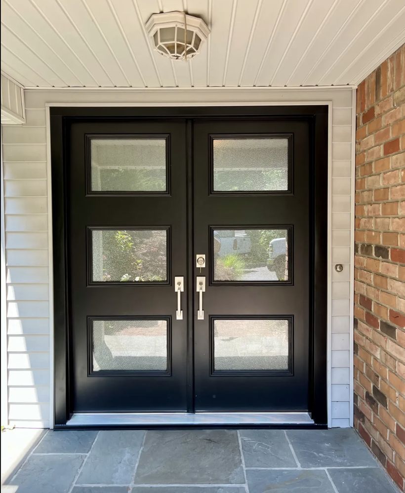 Black double doors with glass panes under a porch ceiling, next to brick and siding.