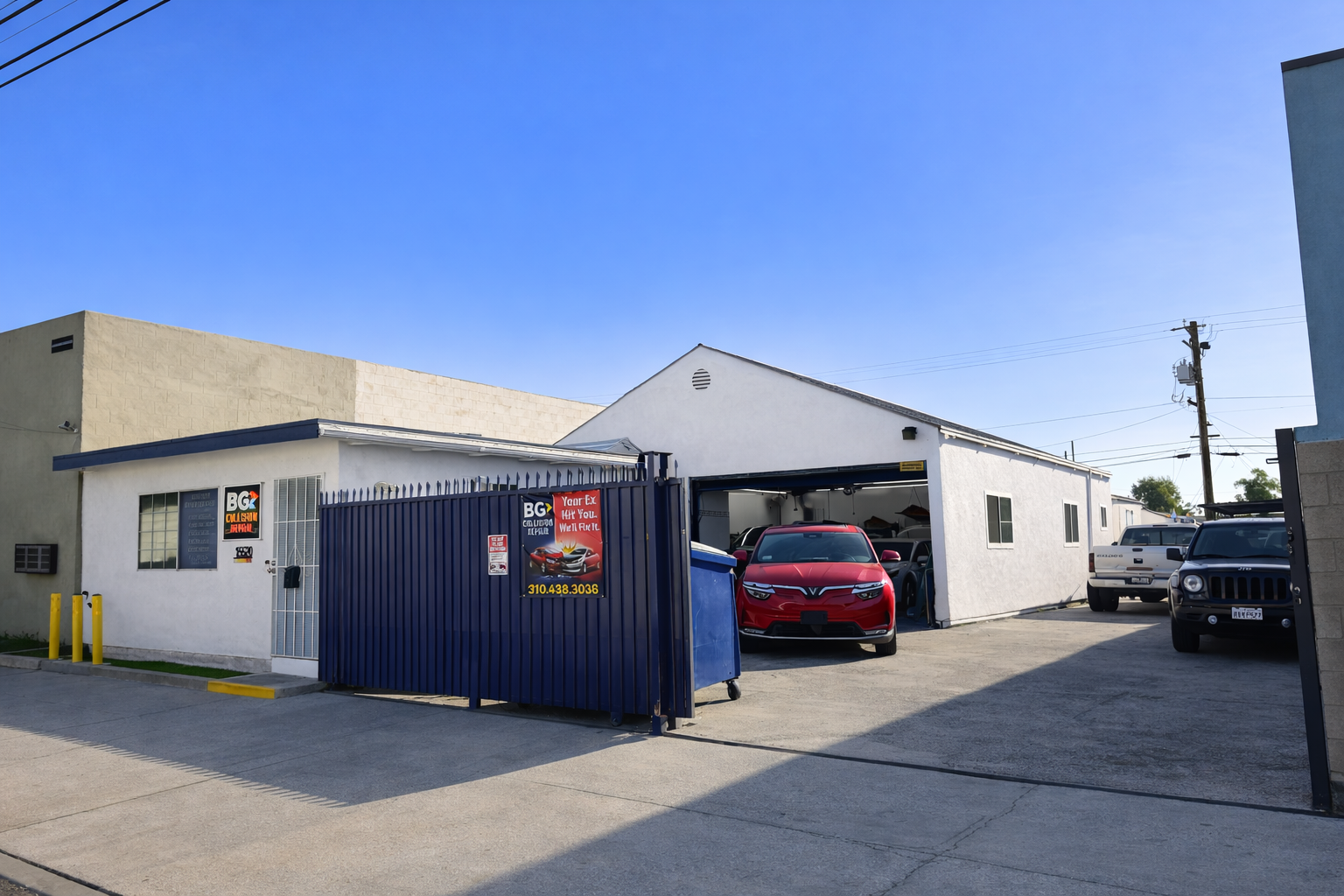 An auto repair shop with a white exterior, a blue fenced entrance, and a red car parked inside the garage bay.