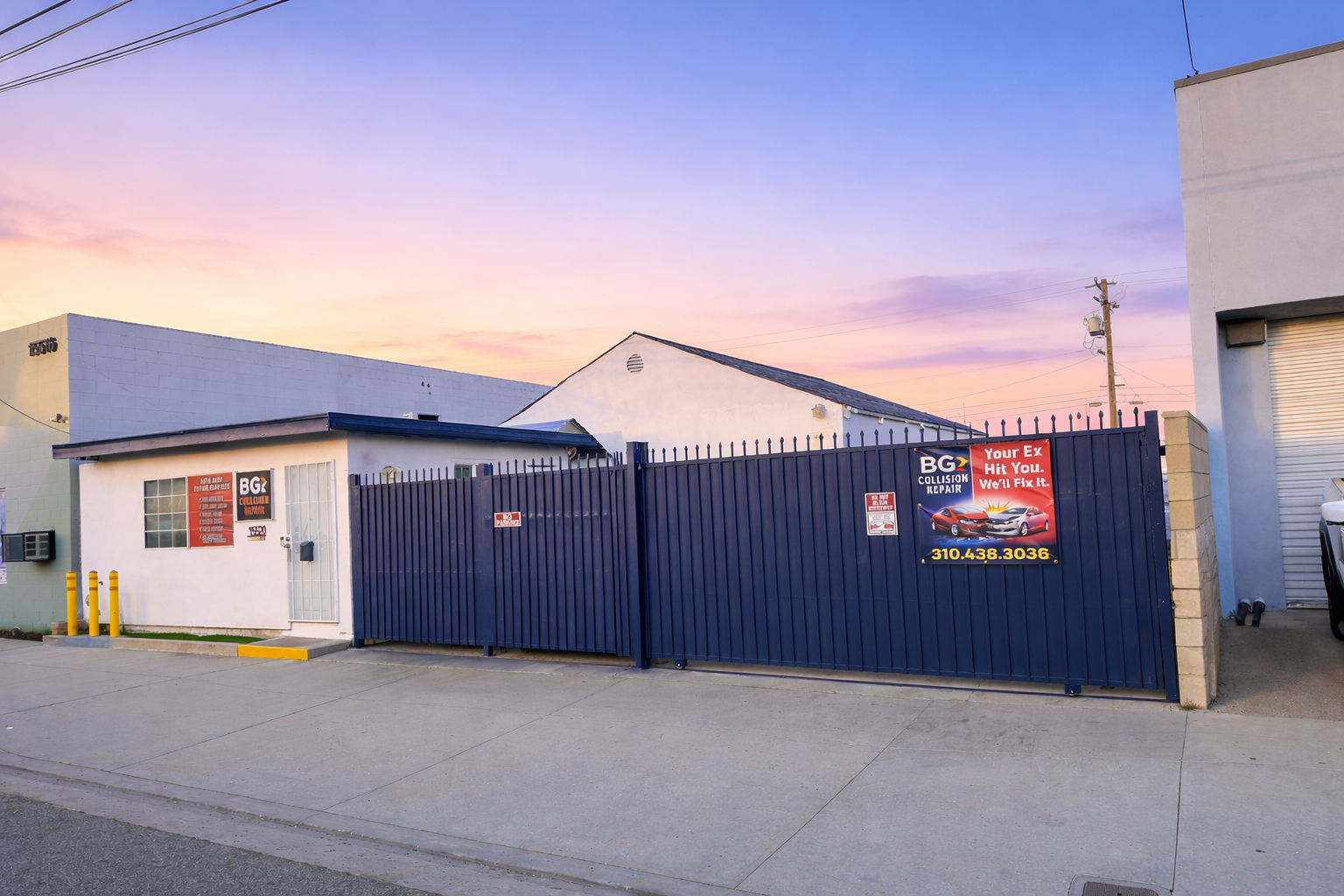 A low-profile white building behind a blue fence with a promotional banner, set against a sunset sky.