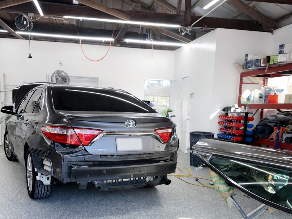 A dark gray car missing its rear bumper sits in a bright, clean auto repair shop with tools and shelving nearby.