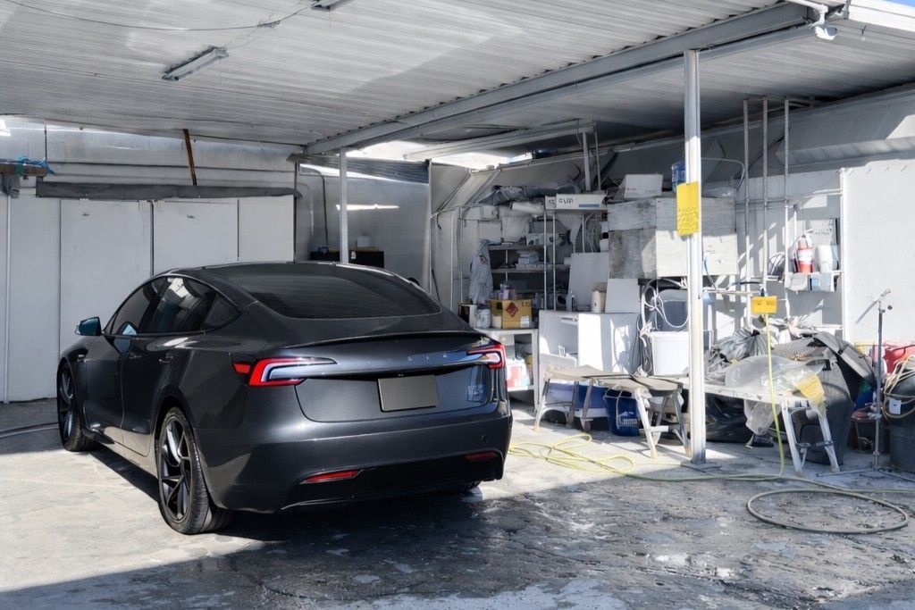 A dark grey Tesla parked under a covered outdoor structure next to a cluttered workspace.