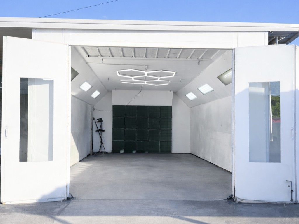 An open, white automotive paint booth with a concrete floor, angular walls, and a distinctive hexagonal LED light fixture.
