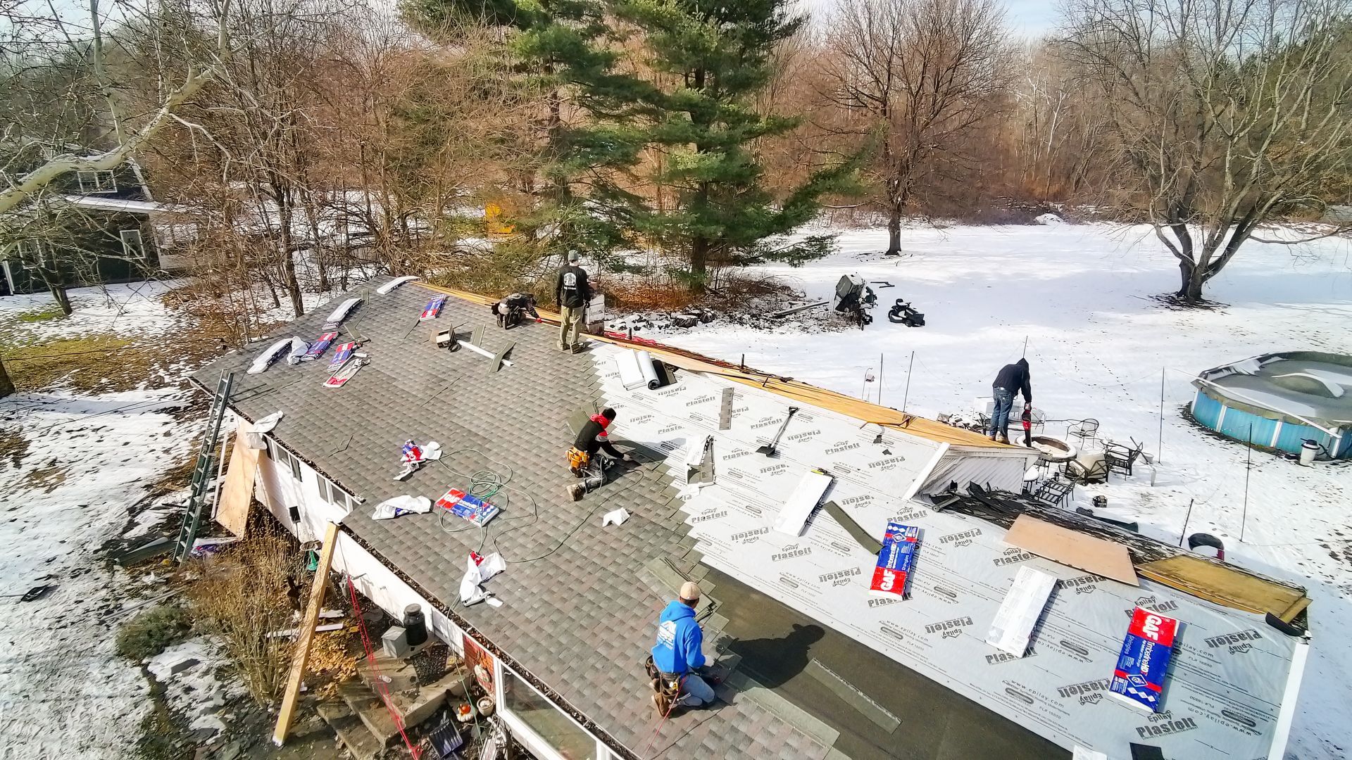 Two roofers removing shingles from a house roof. One roofer wears red, the other, yellow. Orange underlayment is exposed.