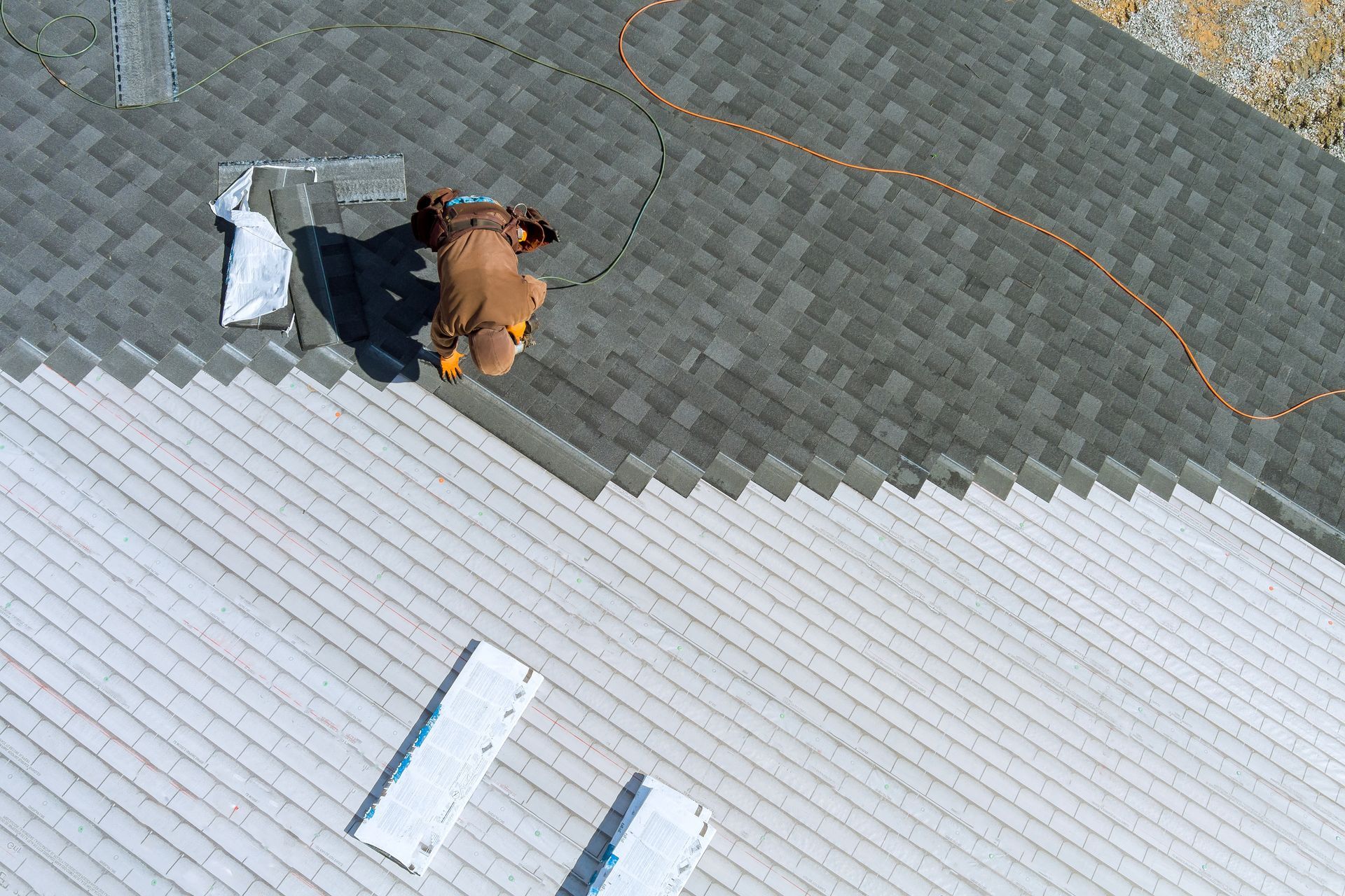 Two workers beside a roof edge, one in an orange shirt with tools and materials on gray shingles